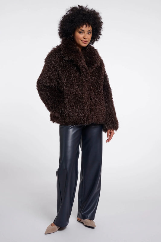A model wearing a dark brown shaggy faux-fur jacket, dark navy wide-leg faux-leather pants, and beige pointed-toe flats, standing against a light gray studio background.