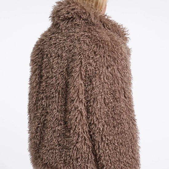 A model shown from the back wearing a shaggy, textured brown faux fur jacket and light beige trousers against a plain white background.