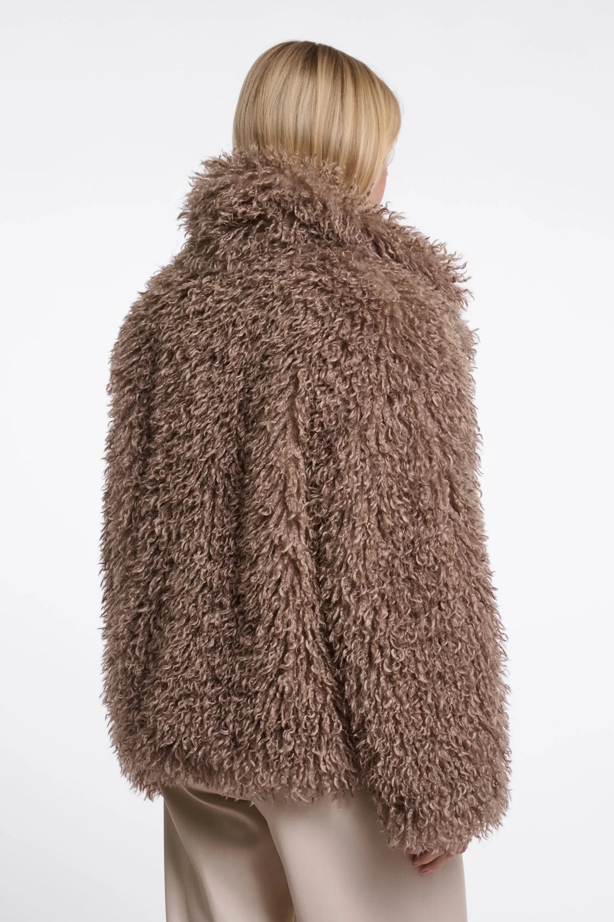 A model shown from the back wearing a shaggy, textured brown faux fur jacket and light beige trousers against a plain white background.