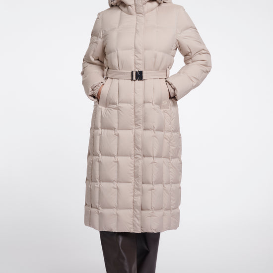 A woman modeling a long, light beige puffer coat with a hood and a matching belt, paired with dark brown pants and boots, against a plain light gray studio background.
