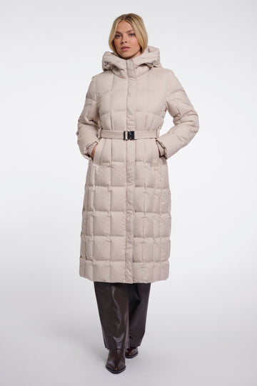 A woman modeling a long, light beige puffer coat with a hood and a matching belt, paired with dark brown pants and boots, against a plain light gray studio background.