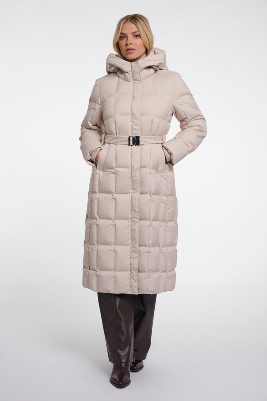 A woman modeling a long, light beige puffer coat with a hood and a matching belt, paired with dark brown pants and boots, against a plain light gray studio background.