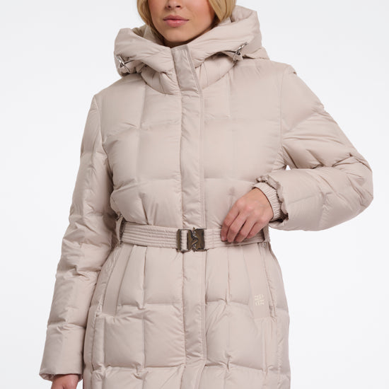 A woman wearing a long, light beige hooded puffer coat with a matching belt and silver buckle, photographed against a plain white studio background.