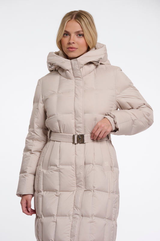 A woman wearing a long, light beige hooded puffer coat with a matching belt and silver buckle, photographed against a plain white studio background.