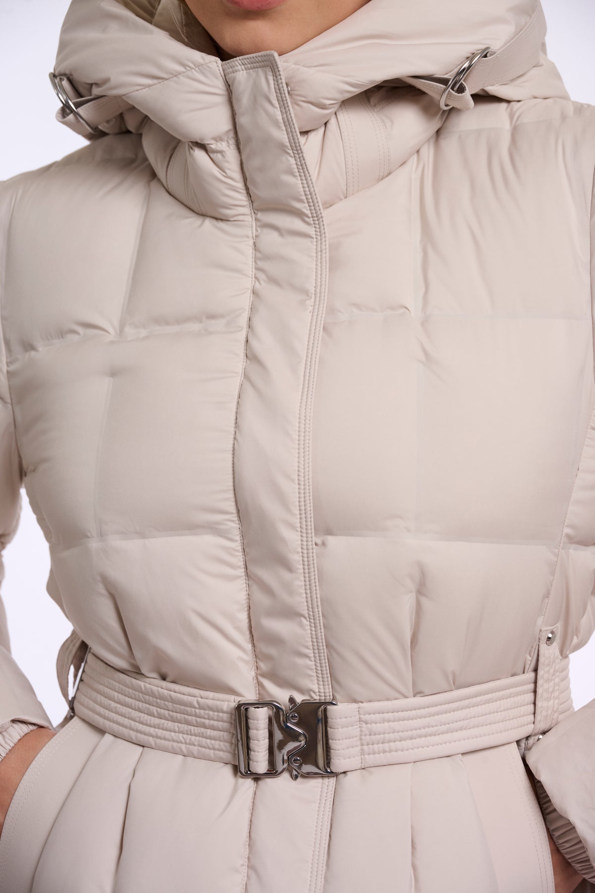 Close-up of a beige hooded puffer jacket with a matching belt and silver buckle, shown on a model.