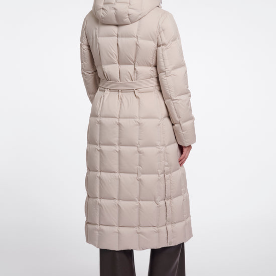 A woman viewed from the back wearing a long, light beige hooded puffer coat with a quilted grid pattern and a matching waist belt, paired with dark wide-leg trousers and black shoes, against a plain light gray studio background.