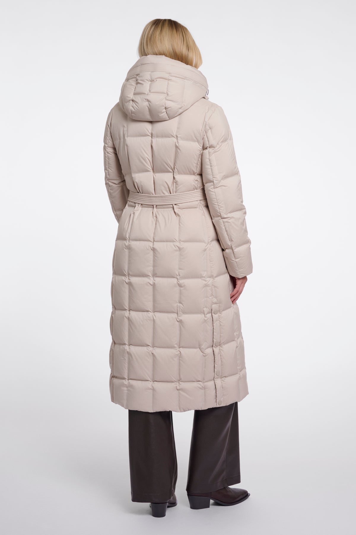 A woman viewed from the back wearing a long, light beige hooded puffer coat with a quilted grid pattern and a matching waist belt, paired with dark wide-leg trousers and black shoes, against a plain light gray studio background.