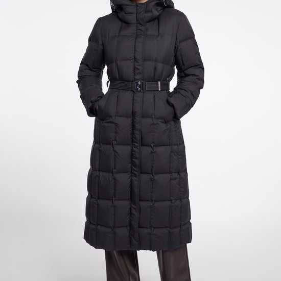 A model wearing a long black puffer coat with a hood and a matching belt, paired with dark brown leather pants, against a plain light gray studio background.