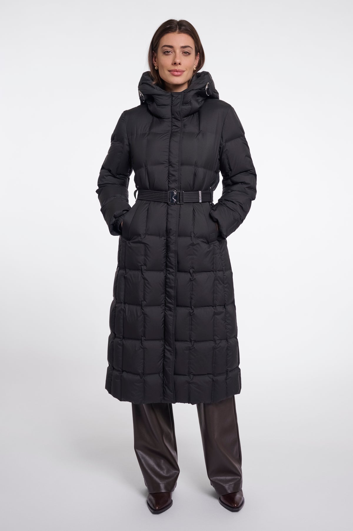 A model wearing a long black puffer coat with a hood and a matching belt, paired with dark brown leather pants, against a plain light gray studio background.