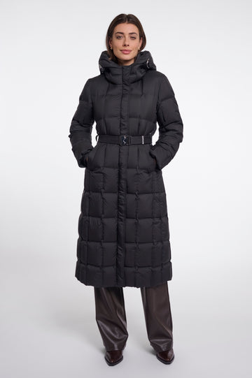 A model wearing a long black puffer coat with a hood and a matching belt, paired with dark brown leather pants, against a plain light gray studio background.