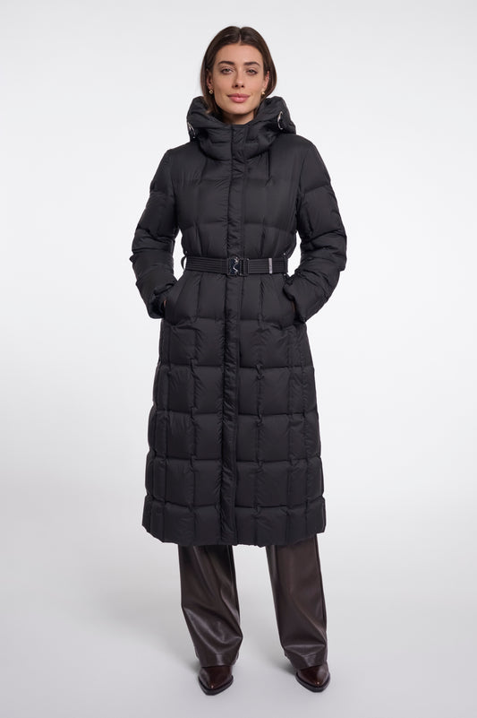 A model wearing a long black puffer coat with a hood and a matching belt, paired with dark brown leather pants, against a plain light gray studio background.
