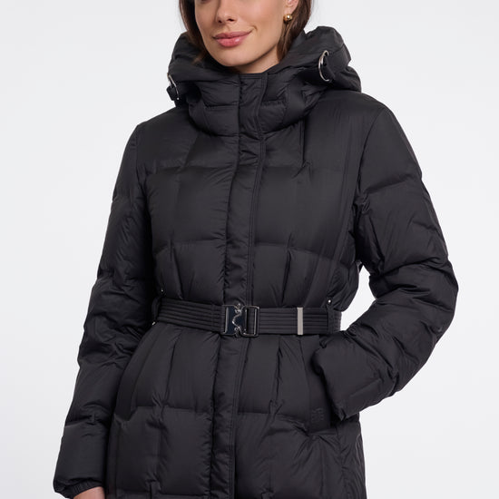 A woman wearing a long black puffer coat with a hood and a matching belt, photographed against a plain light gray studio background.