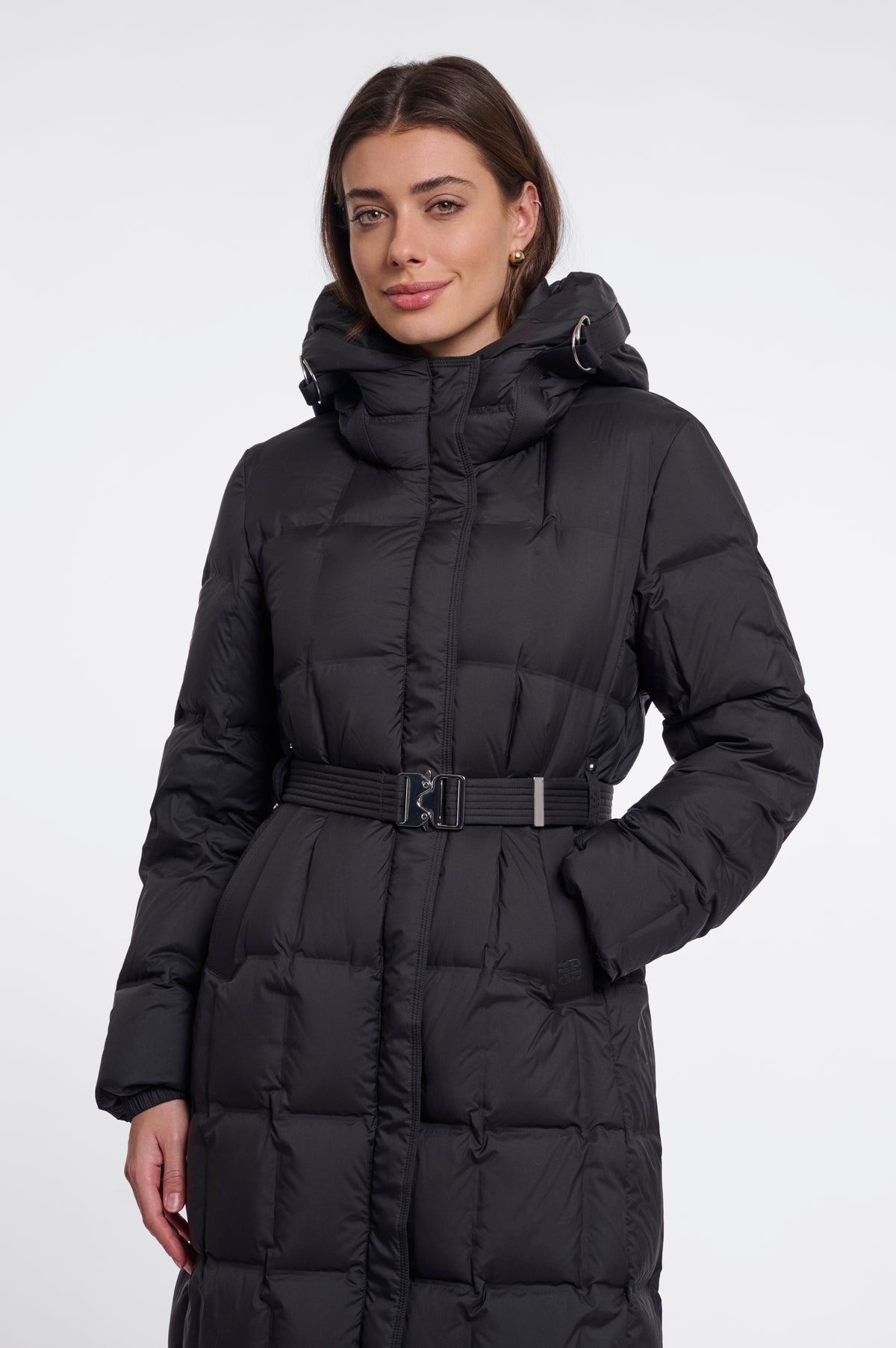 A woman wearing a long black puffer coat with a hood and a matching belt, photographed against a plain light gray studio background.
