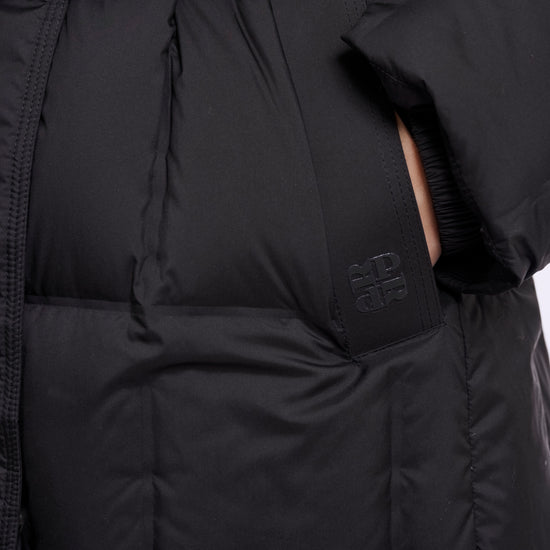 Close-up of a black puffer jacket with horizontal quilting, a matching black belt with a silver buckle, and a small tonal logo on the side pocket.