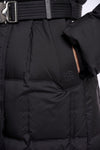 Close-up of a black puffer jacket with horizontal quilting, a matching black belt with a silver buckle, and a small tonal logo on the side pocket.