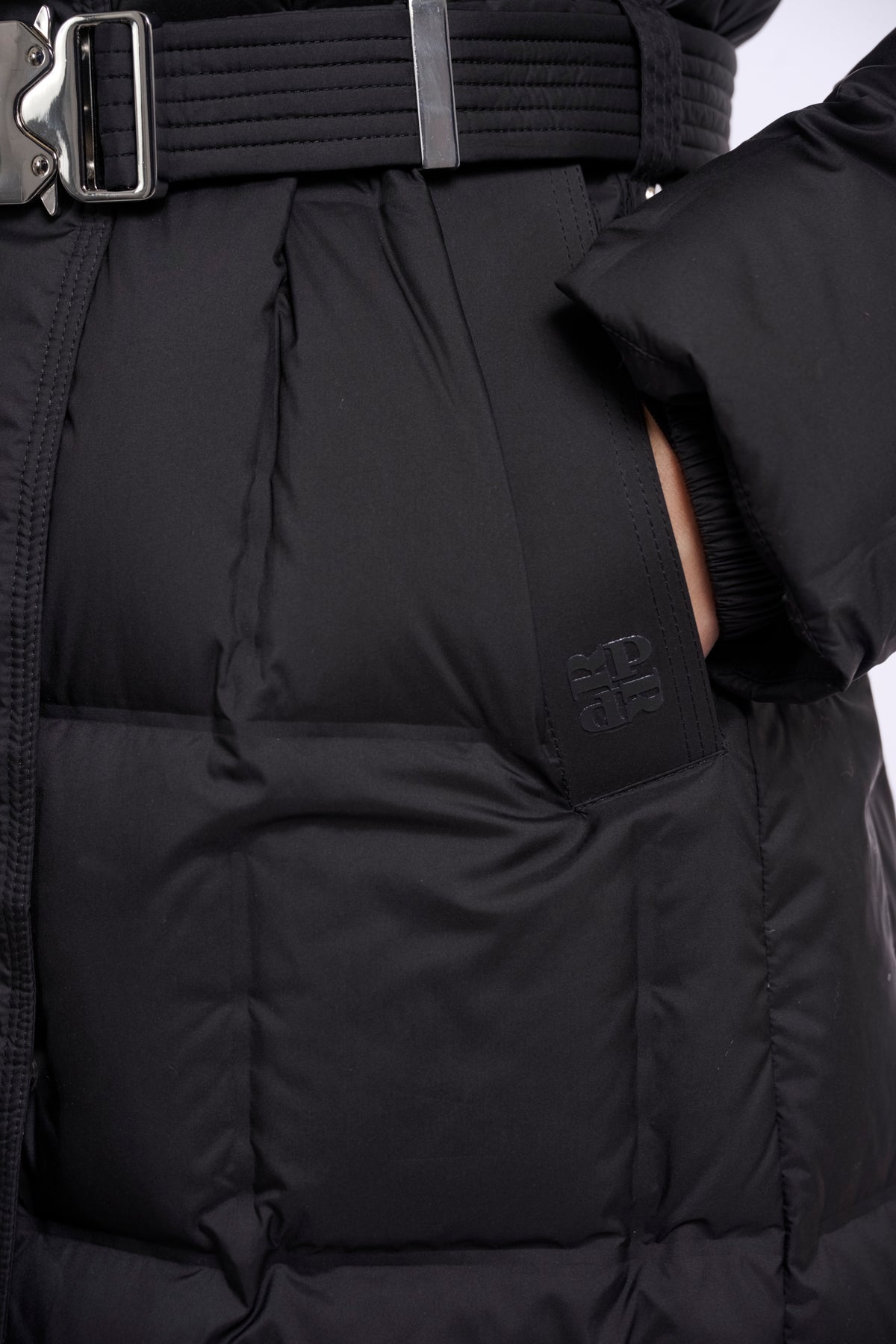 Close-up of a black puffer jacket with horizontal quilting, a matching black belt with a silver buckle, and a small tonal logo on the side pocket.