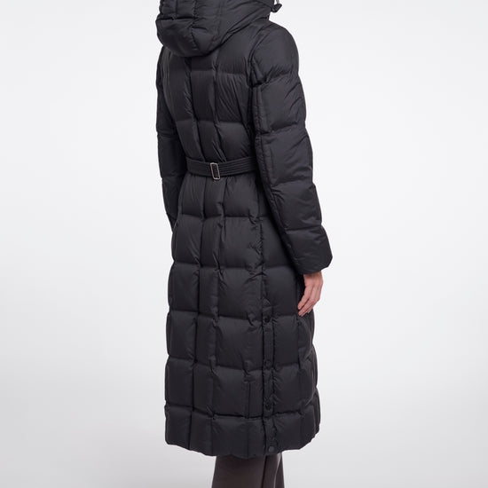 A model shown from the back wearing a long black hooded puffer coat with a belted waist, paired with dark wide-leg trousers, against a plain light gray studio background.