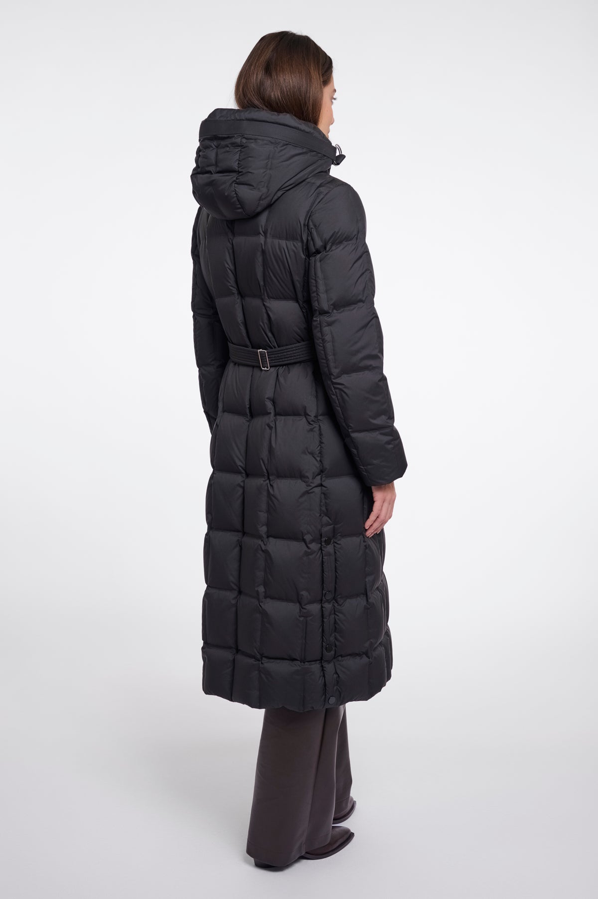 A model shown from the back wearing a long black hooded puffer coat with a belted waist, paired with dark wide-leg trousers, against a plain light gray studio background.