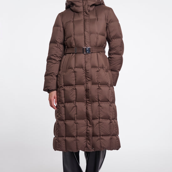 A model wearing a long, brown, quilted puffer coat with a matching belt and hood, paired with black pants, against a plain light gray studio background.