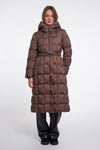 A model wearing a long, brown, quilted puffer coat with a matching belt and hood, paired with black pants, against a plain light gray studio background.