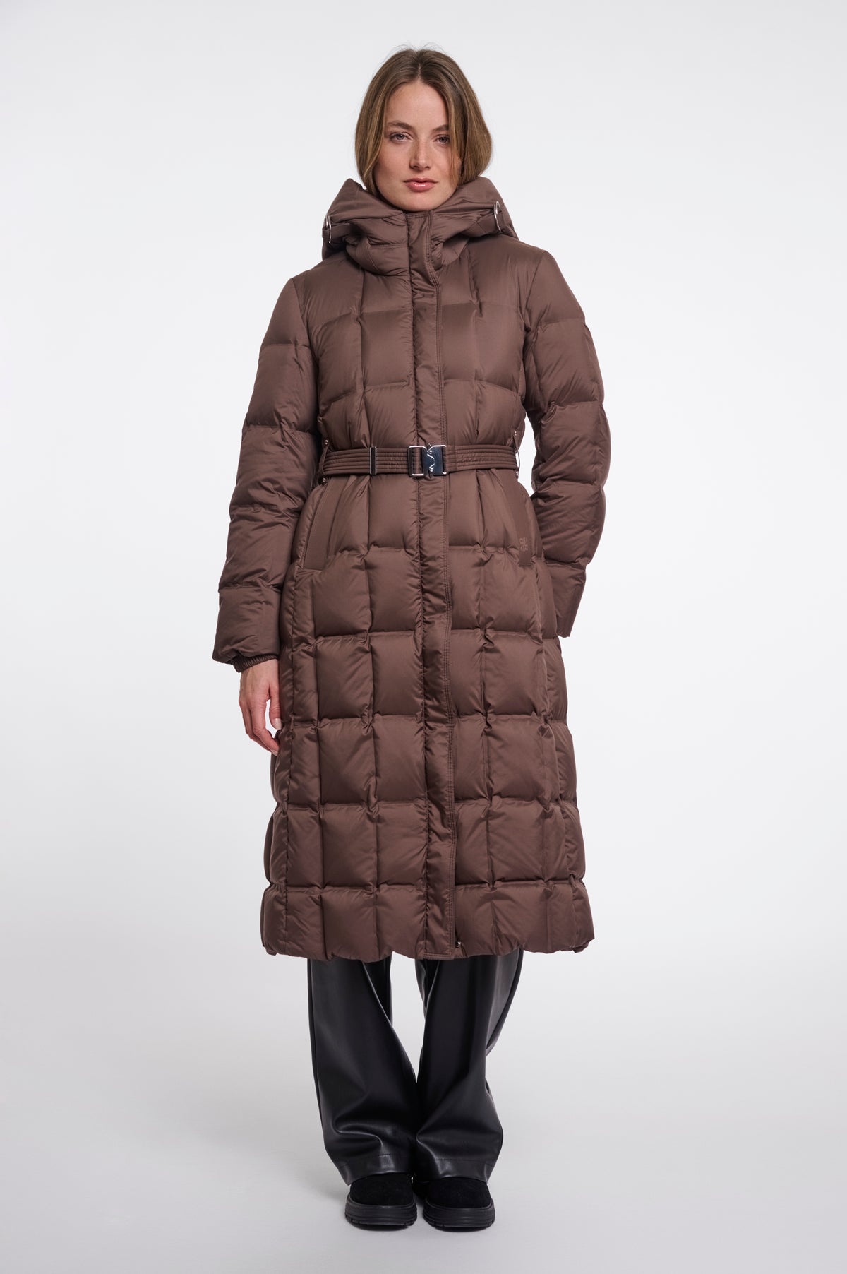 A model wearing a long, brown, quilted puffer coat with a matching belt and hood, paired with black pants, against a plain light gray studio background.