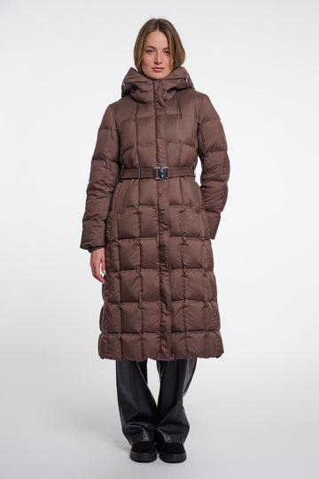 A model wearing a long, brown, quilted puffer coat with a matching belt and hood, paired with black pants, against a plain light gray studio background.