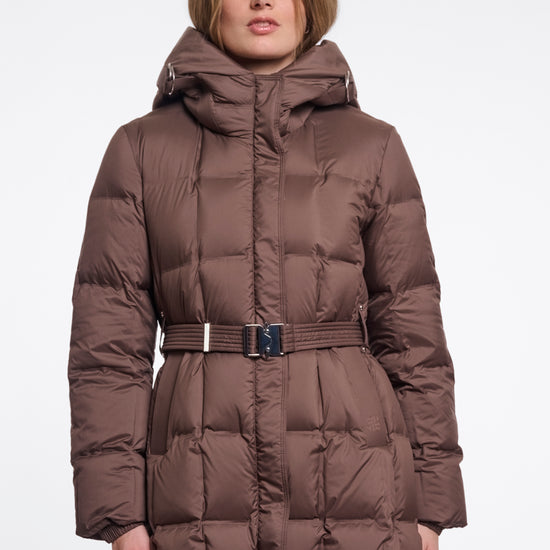 A woman wearing a long brown puffer coat with a hood and a matching belt, photographed against a plain light gray studio background.