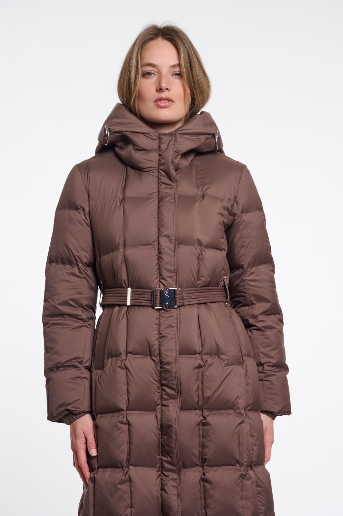 A woman wearing a long brown puffer coat with a hood and a matching belt, photographed against a plain light gray studio background.