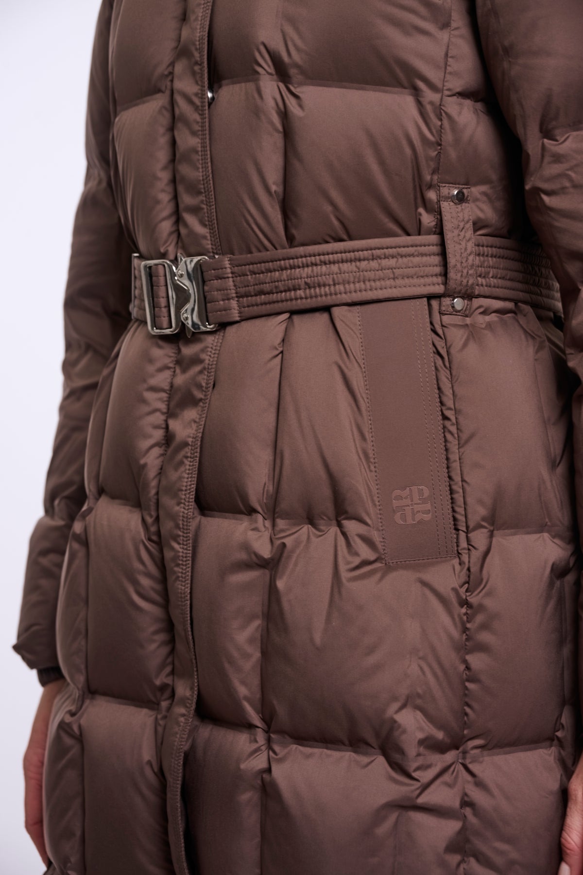 A close-up of a brown puffer coat with horizontal quilting, a matching belt with a silver buckle, and a small tonal logo patch on the side.