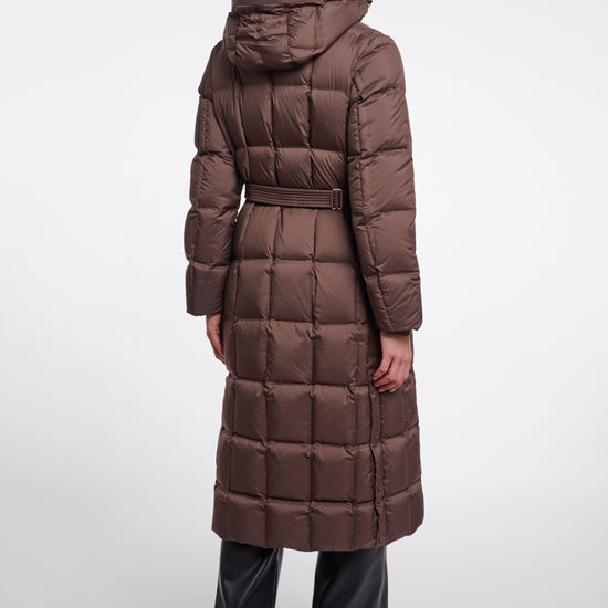 A model viewed from the back wearing a long brown puffer coat with a hood and waist belt, paired with black pants, against a light gray studio background.