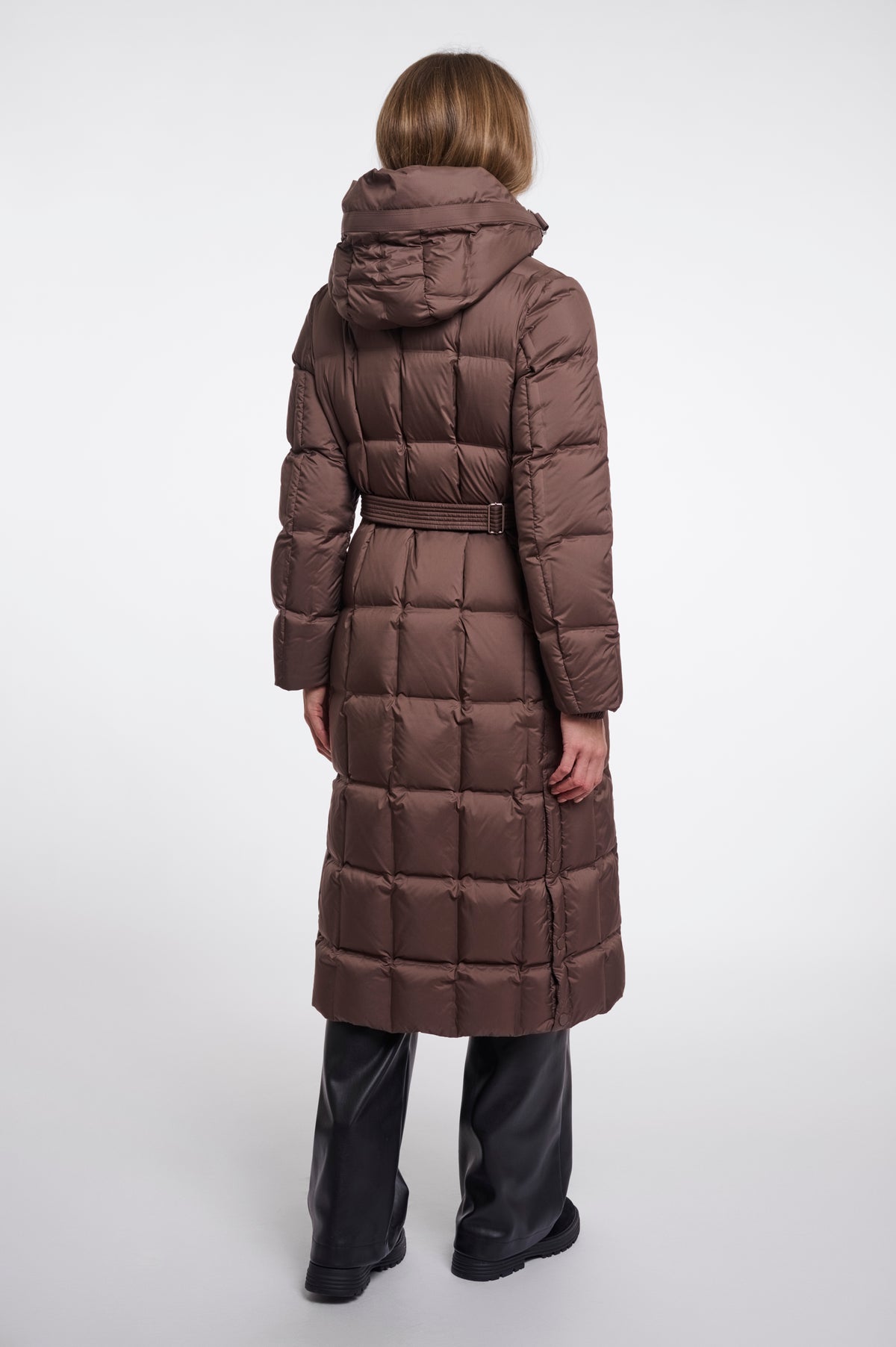A model viewed from the back wearing a long brown puffer coat with a hood and waist belt, paired with black pants, against a light gray studio background.