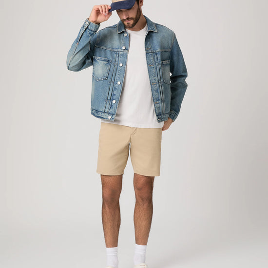 Man wearing a denim jacket, white t-shirt, beige shorts, and white sneakers on a plain background from Paige