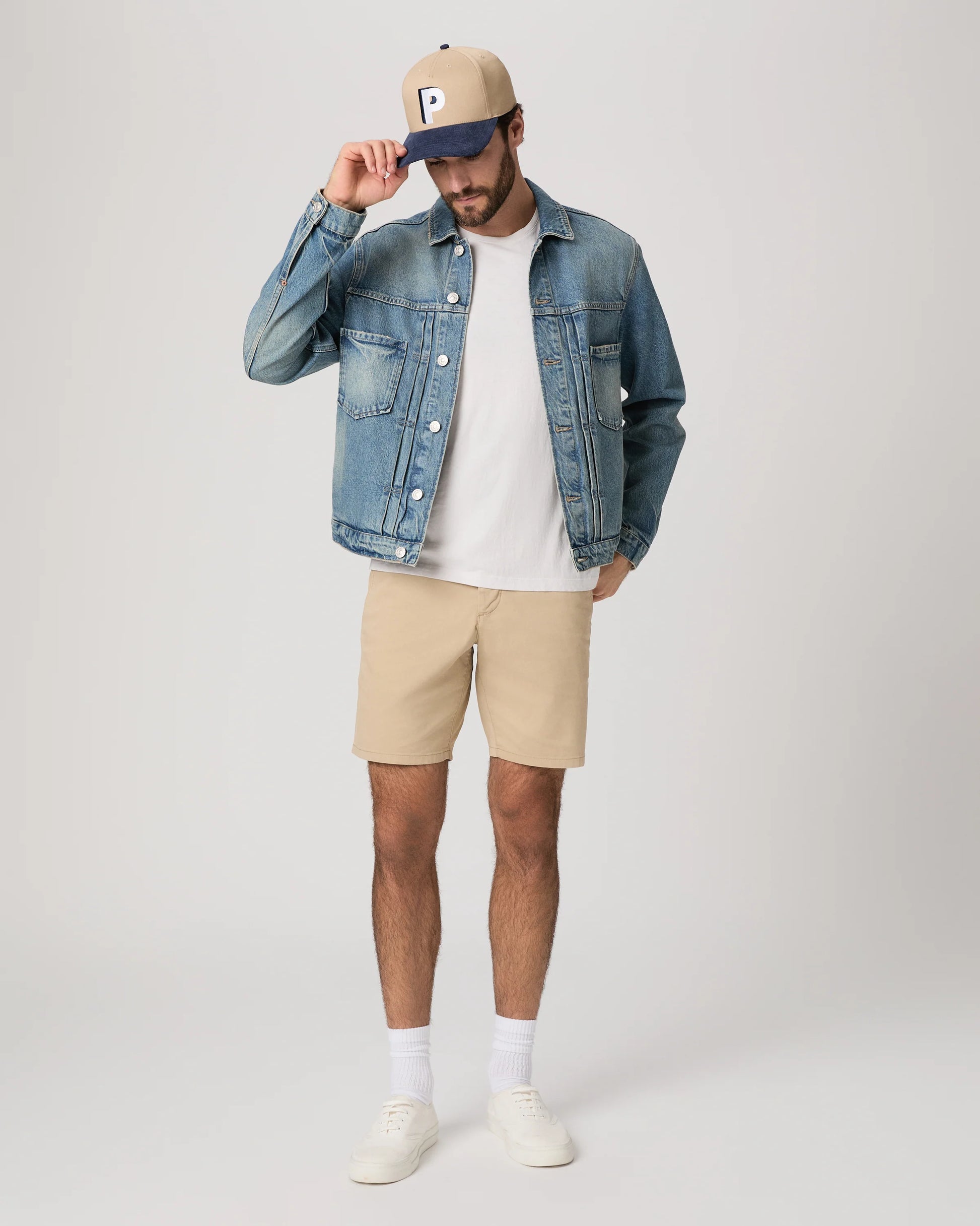 Man wearing a denim jacket, white t-shirt, beige shorts, and white sneakers on a plain background from Paige