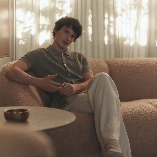Person sitting on a couch in a softly lit room with curtains wearing beige paige pants
