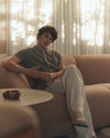 Person sitting on a couch in a softly lit room with curtains wearing beige paige pants