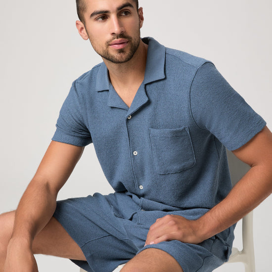 Man sat on a chair wearing a blue paige shirt on a white background
