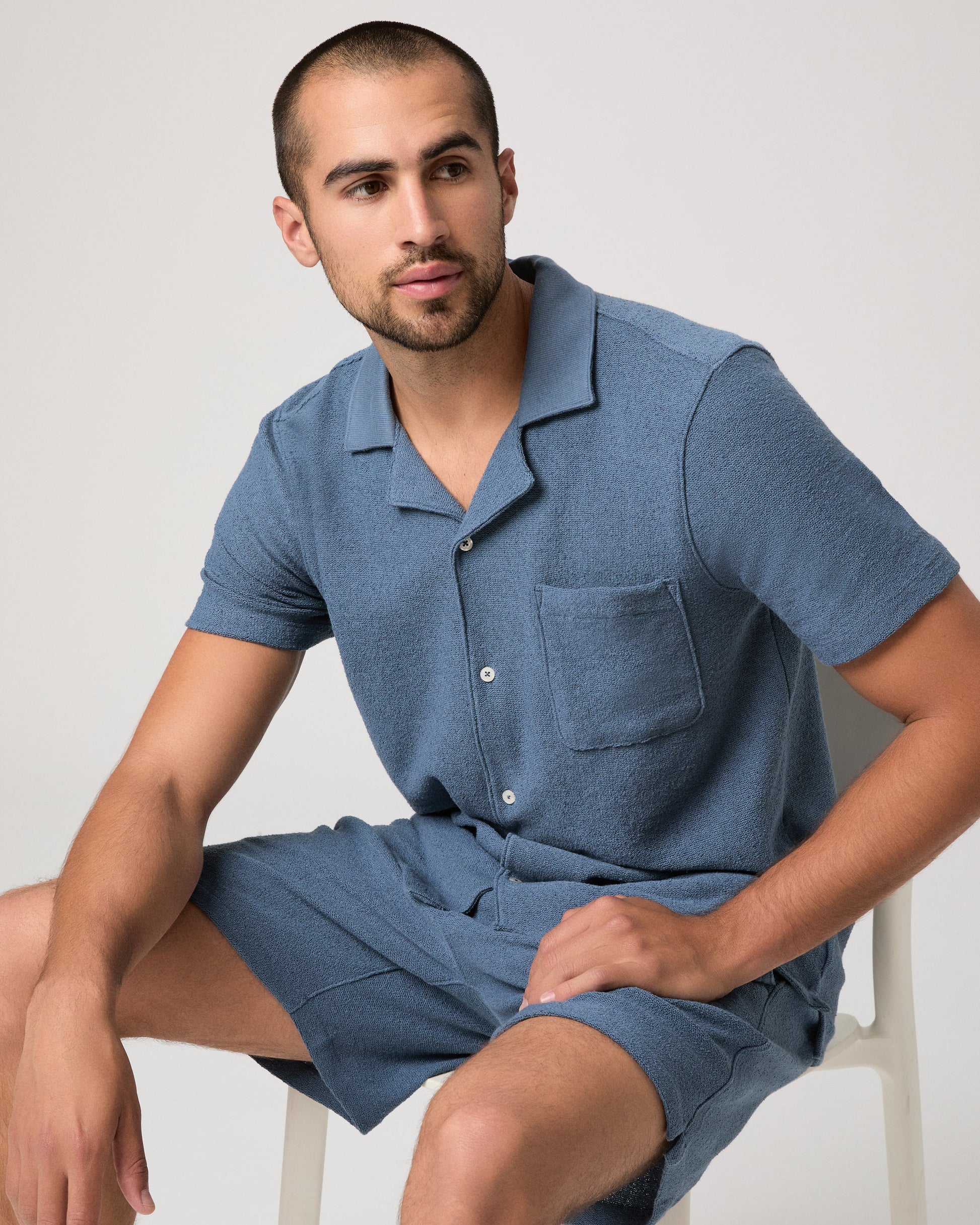 Man sat on a chair wearing a blue paige shirt on a white background