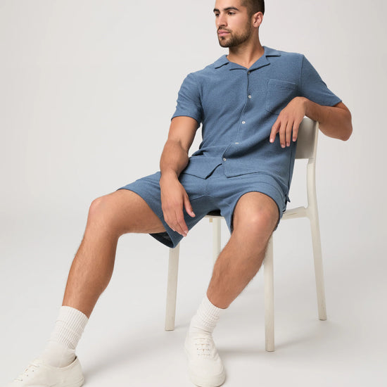 Man sat on a chair wearing a blue paige shirt and matching shorts on a white background