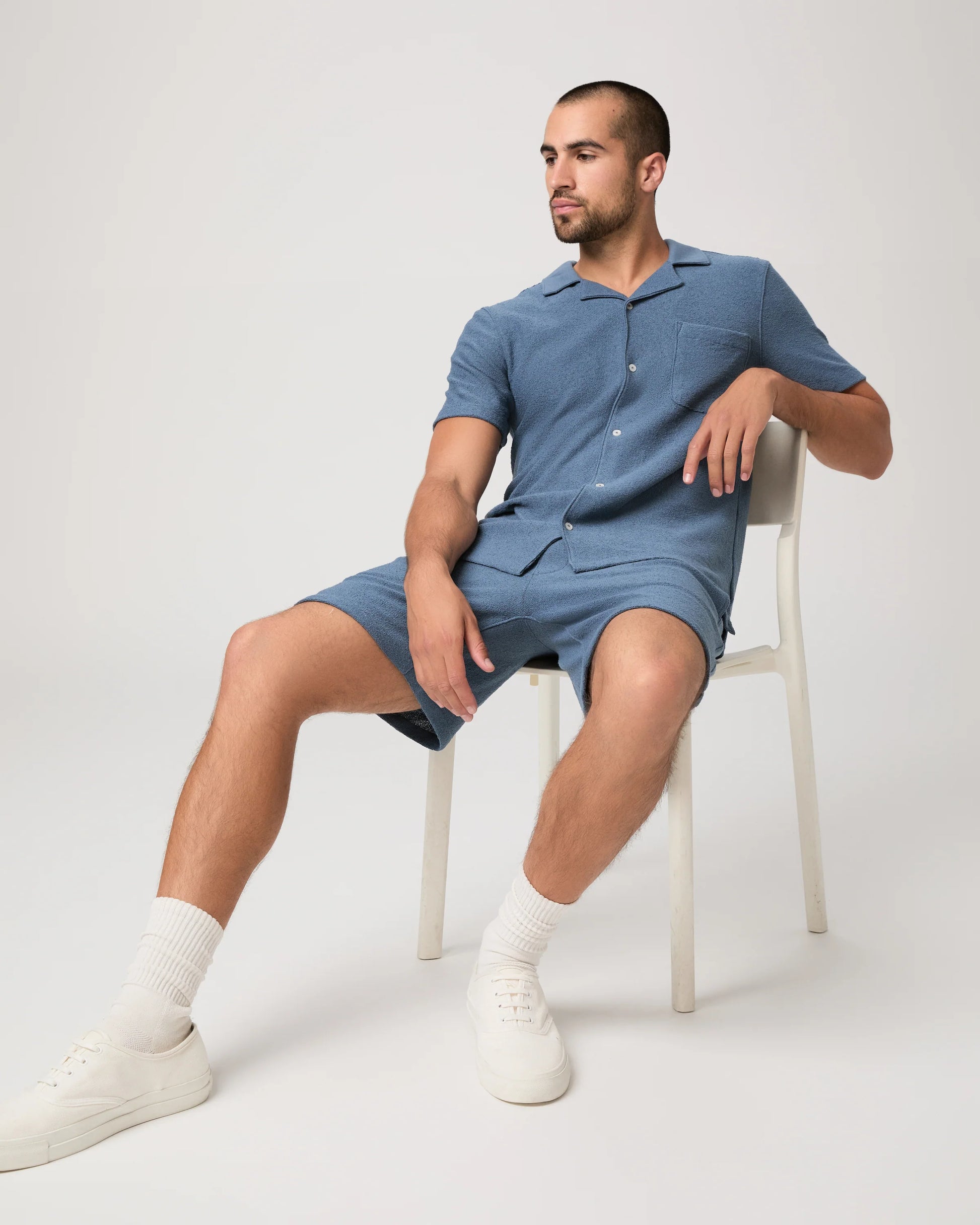 Man sat on a chair wearing a blue paige shirt and matching shorts on a white background