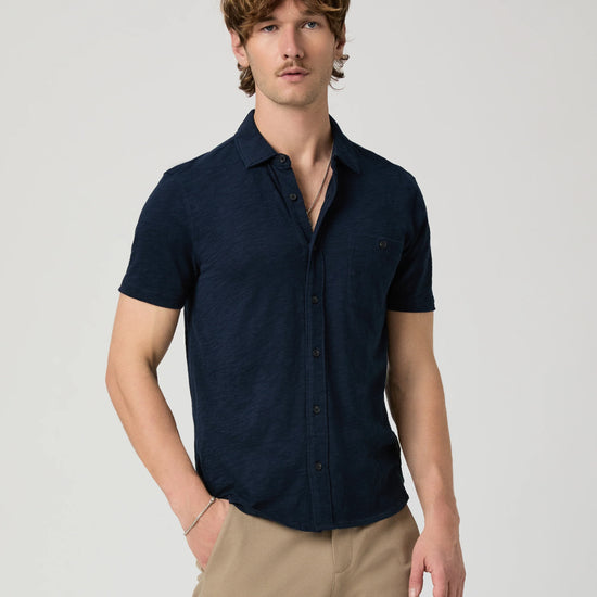Man wearing a navy short-sleeve PAIGE shirt and beige pants on a light gray background