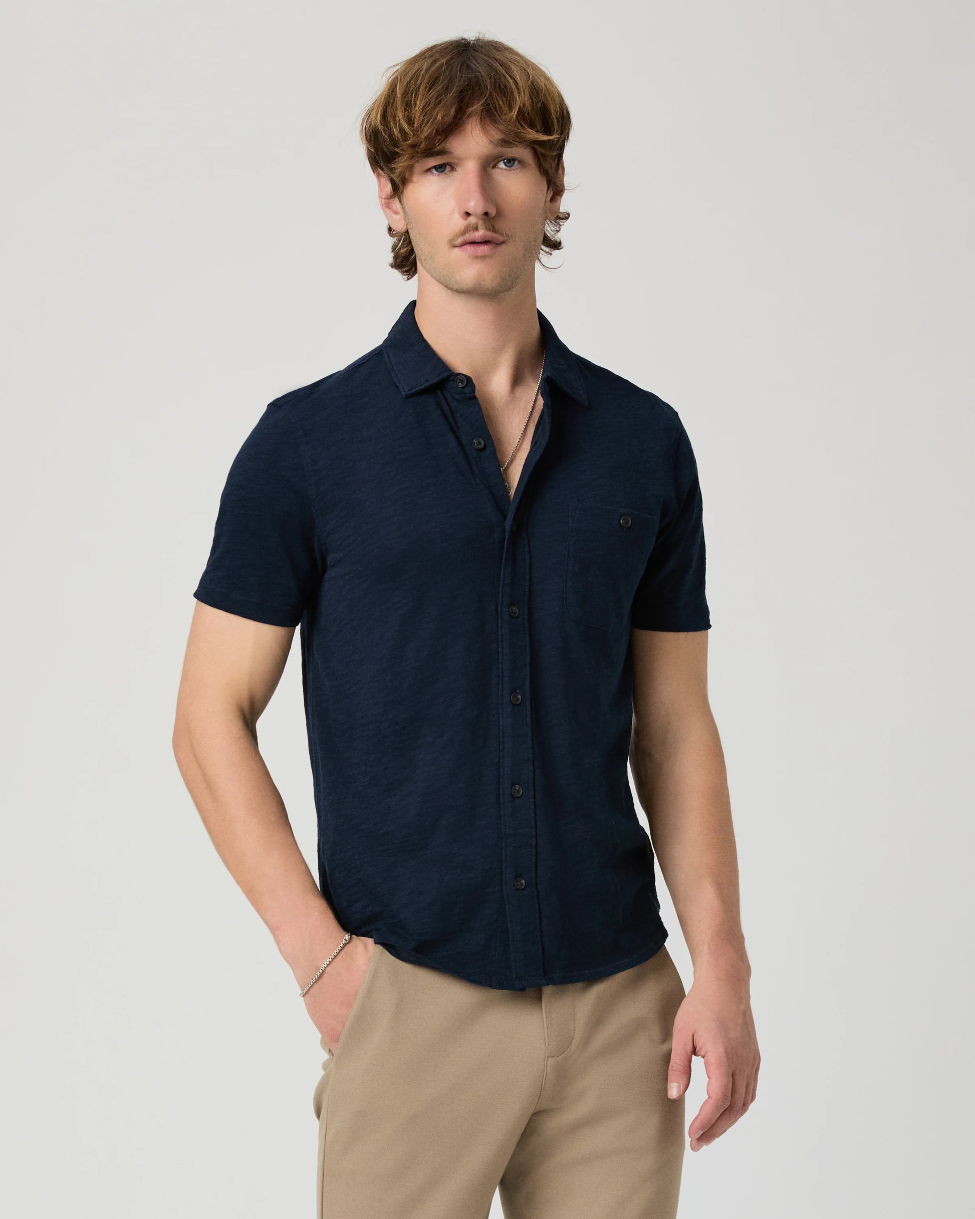 Man wearing a navy short-sleeve PAIGE shirt and beige pants on a light gray background
