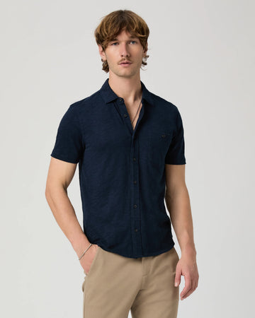 Man wearing a navy short-sleeve PAIGE shirt and beige pants on a light gray background