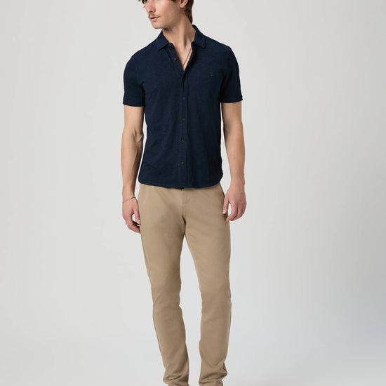 Man wearing a navy short-sleeve PAIGE shirt and beige pants on a light gray background another angle 