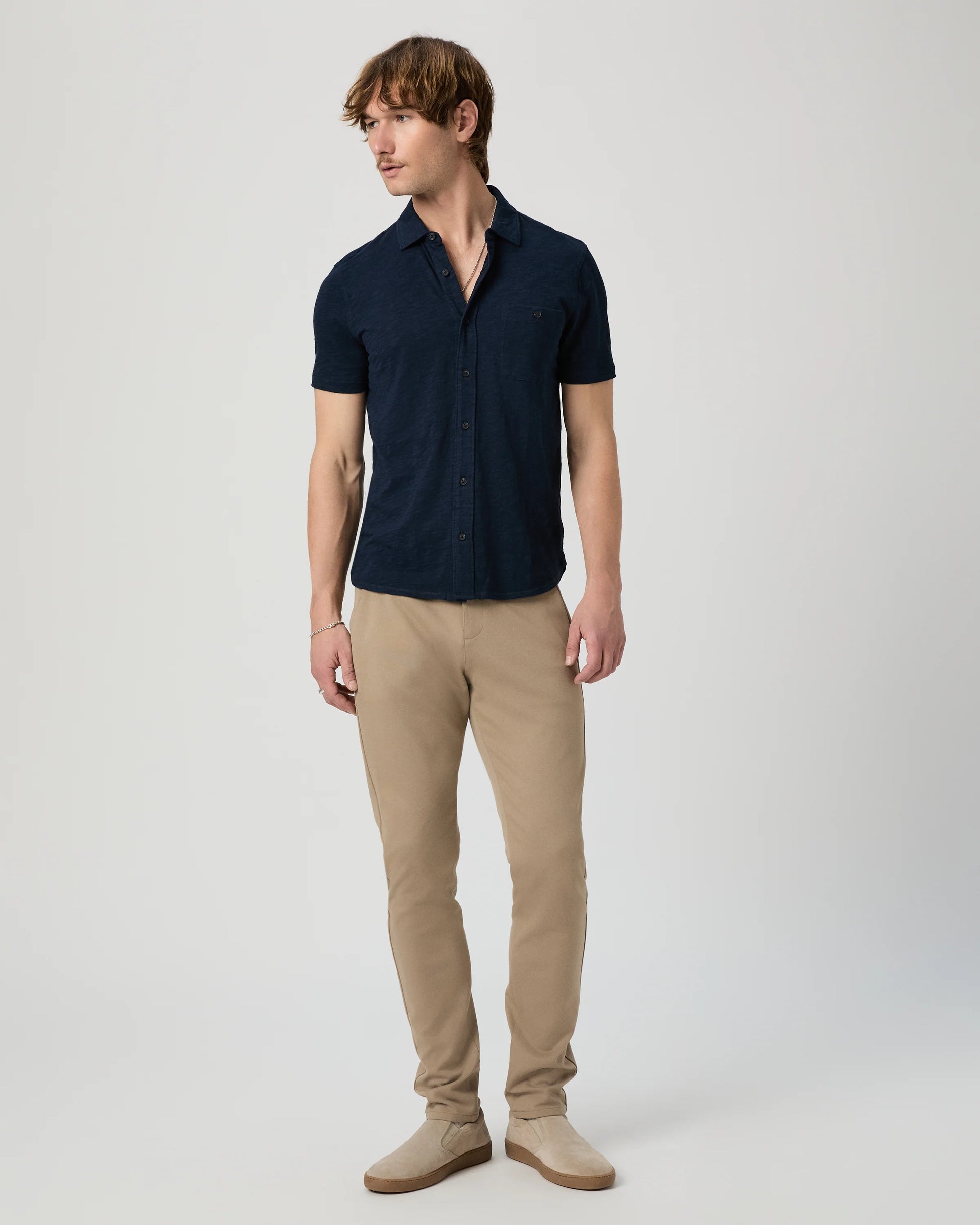 Man wearing a navy short-sleeve PAIGE shirt and beige pants on a light gray background another angle 