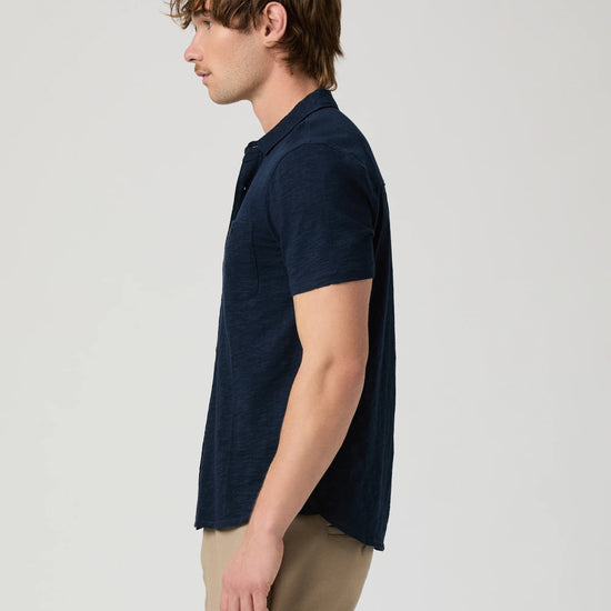 Man wearing a navy short-sleeve PAIGE shirt and beige pants on a light gray background side view