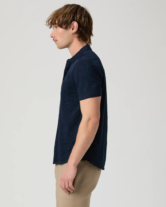 Man wearing a navy short-sleeve PAIGE shirt and beige pants on a light gray background side view