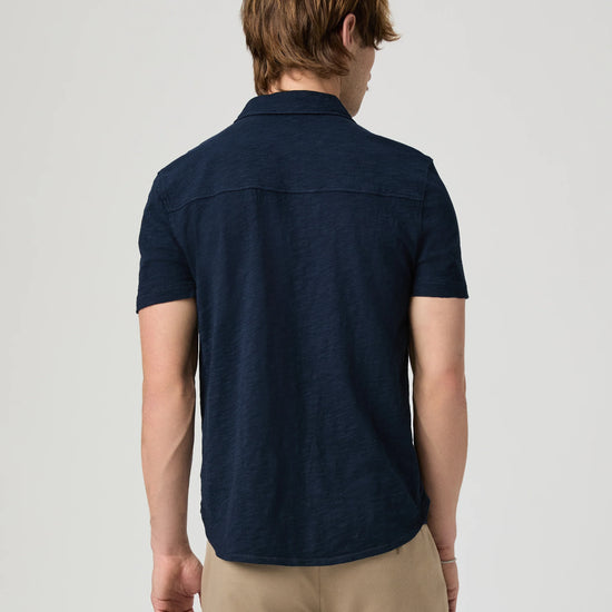 Man wearing a navy short-sleeve PAIGE shirt and beige pants on a light gray background rear view