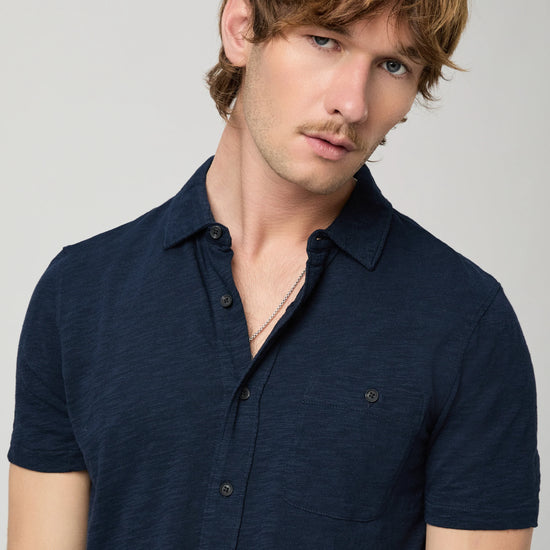 Man wearing a navy short-sleeve PAIGE shirt and beige pants on a light gray background close up 