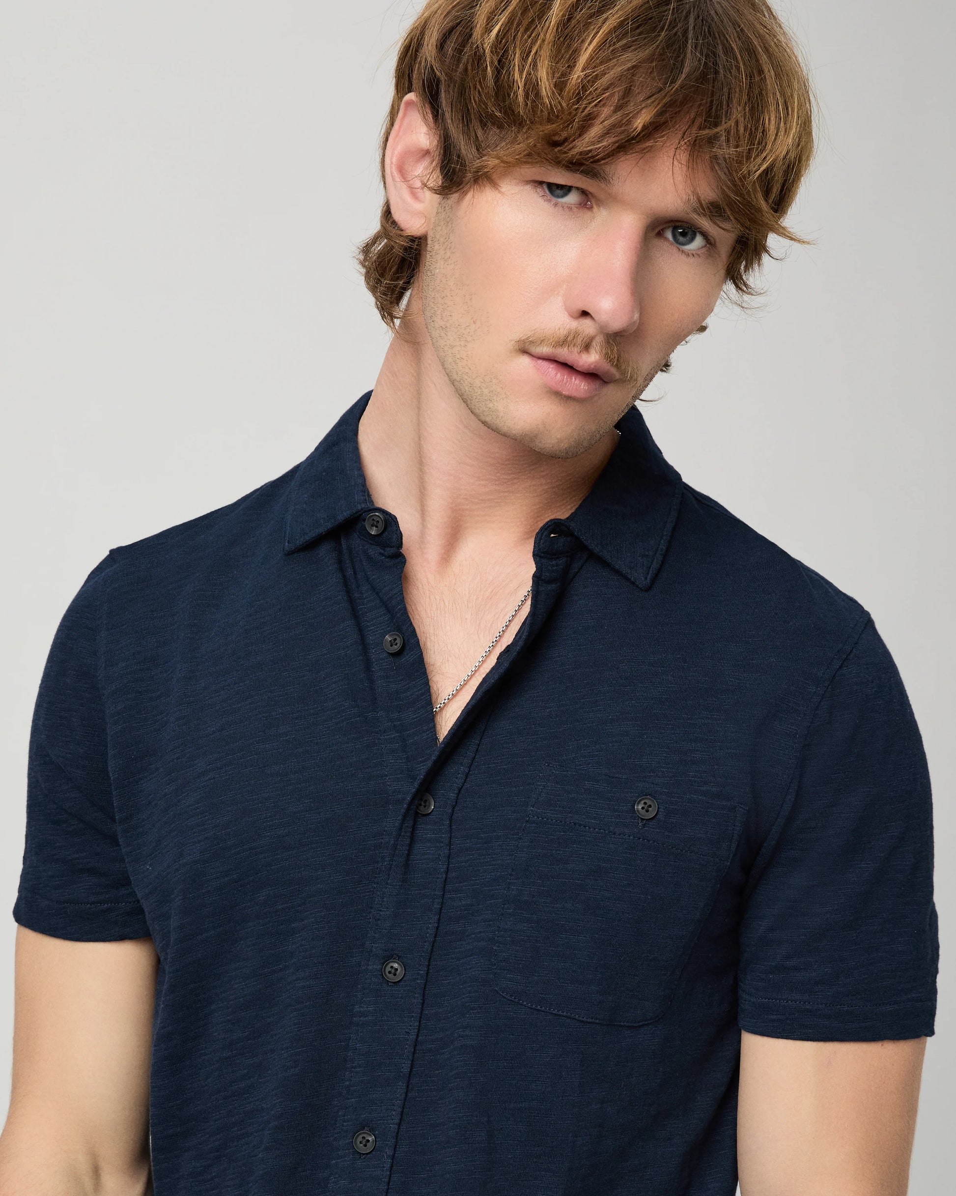 Man wearing a navy short-sleeve PAIGE shirt and beige pants on a light gray background close up 