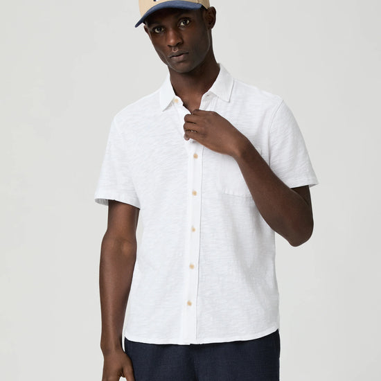 Man wearing a white short-sleeve paige shirt and beige cap with a logo on a plain background